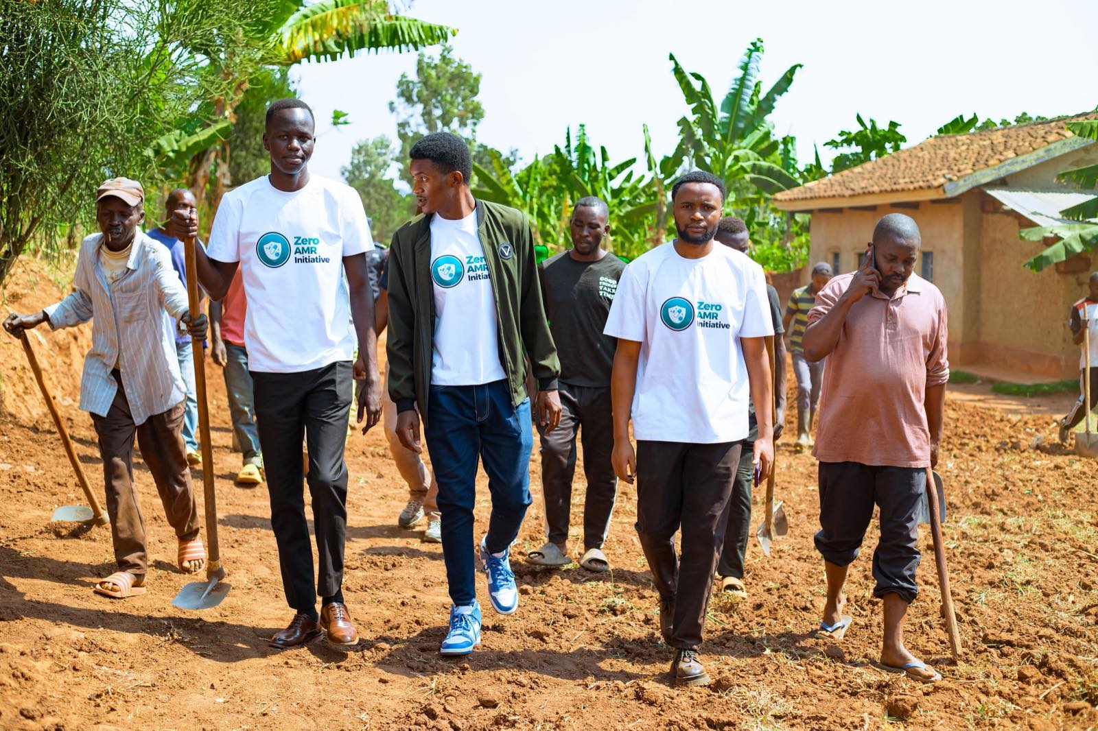Zero AMR Initiative team members in branded white shirts walking along a rural path alongside community members carrying hoes