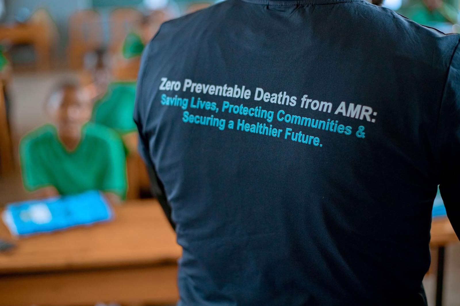 The back of a Zero AMR Initiative shirt reading Zero Preventable Deaths from AMR: Saving Lives, Protecting Communities and Securing a Healthier Future, with a student seated behind